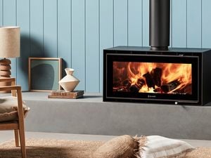 Black freestanding wood heater with burning logs against a light blue wall, next to a log holder with a rug and cushion on the floor.