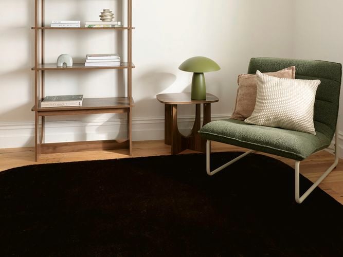 Warm-toned living room with a textured modular sofa styled with rust and olive cushions, a wooden coffee table on a mauve rug, and sunlight streaming through a large window beside indoor plants and timber shelving.