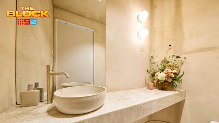 A furnished bathroom with a basin, tap, vase with flowers in it on a marble benchtop and lights on the wall.