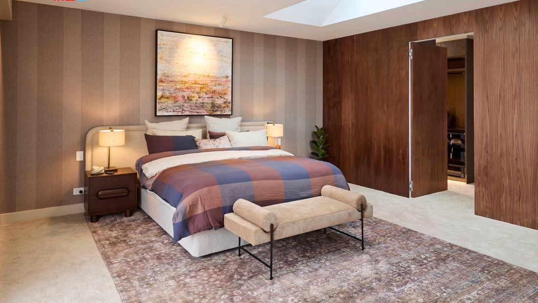 A modern bedroom with a large bed featuring a blue and brown checkered blanket, flanked by two bedside tables with lamps. A large abstract painting hangs above the bed. The room has beige carpet, wooden panel walls, and an upholstered bench at the foot of the bed. A doorway on the right leads to another room. The ceiling includes recessed lighting and a skylight. 