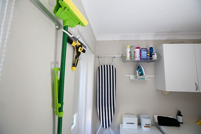 Floor mop, ironing board and wire basket of products hanging from wall mounted rail in a laundry