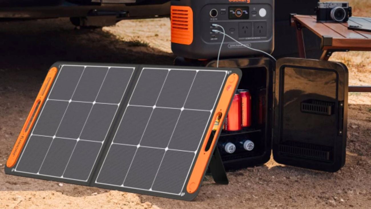 Portable solar panels connected to a Jackery power station, placed near a vehicle, with a mini fridge and camera on a table nearby.