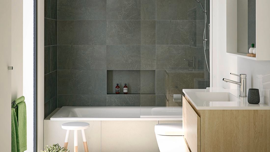 A modern bathroom with grey tiles, a white bathtub with shower, toilet, and a wooden vanity with a white basin. Three green towels hang on a rack to the left next to a white and timber stool.