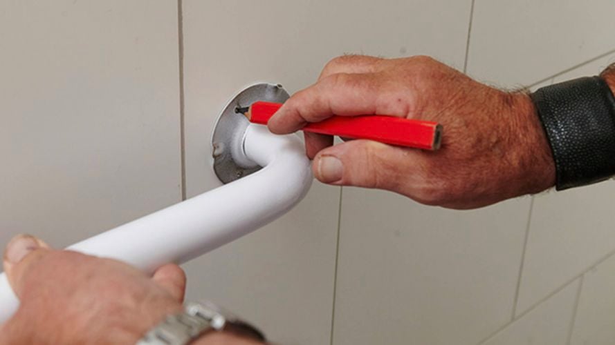  Person marking a wall with a red pencil while positioning a white grab bar on a metal mounting plate.