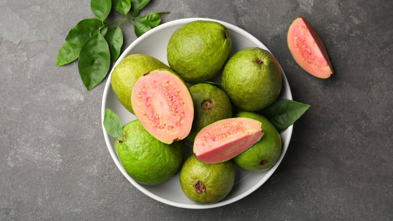 An image of fresh whole and cut guava fruits displayed in a white bowl on a grey textured table