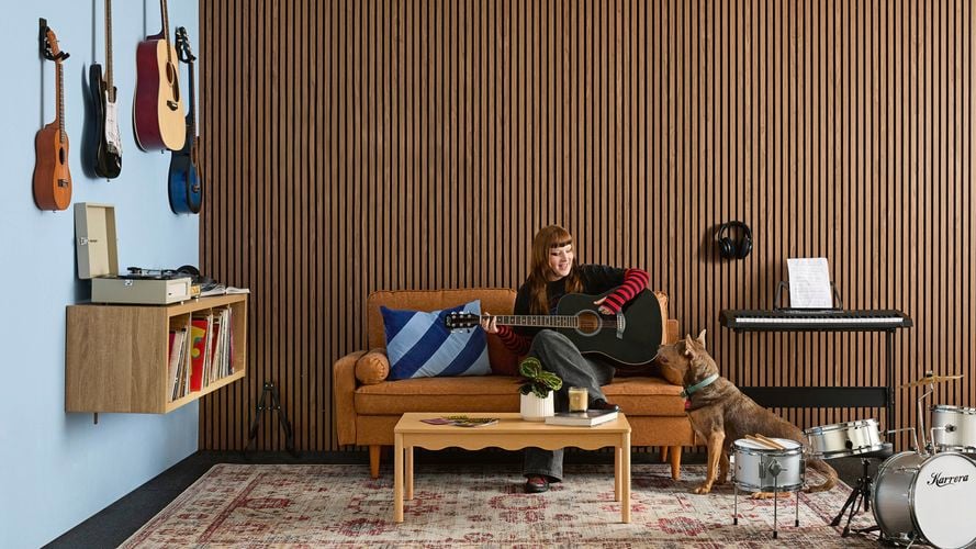 A music room with acoustic wall panelling, rubber floor matting, a tan sofa, drum kit, record player on floating shelves, wall-mounted guitars and a keyboard 