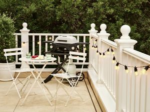 Deck with white balustrade, string lights, a gas barbecue, and a small outdoor dining set surrounded by greenery.