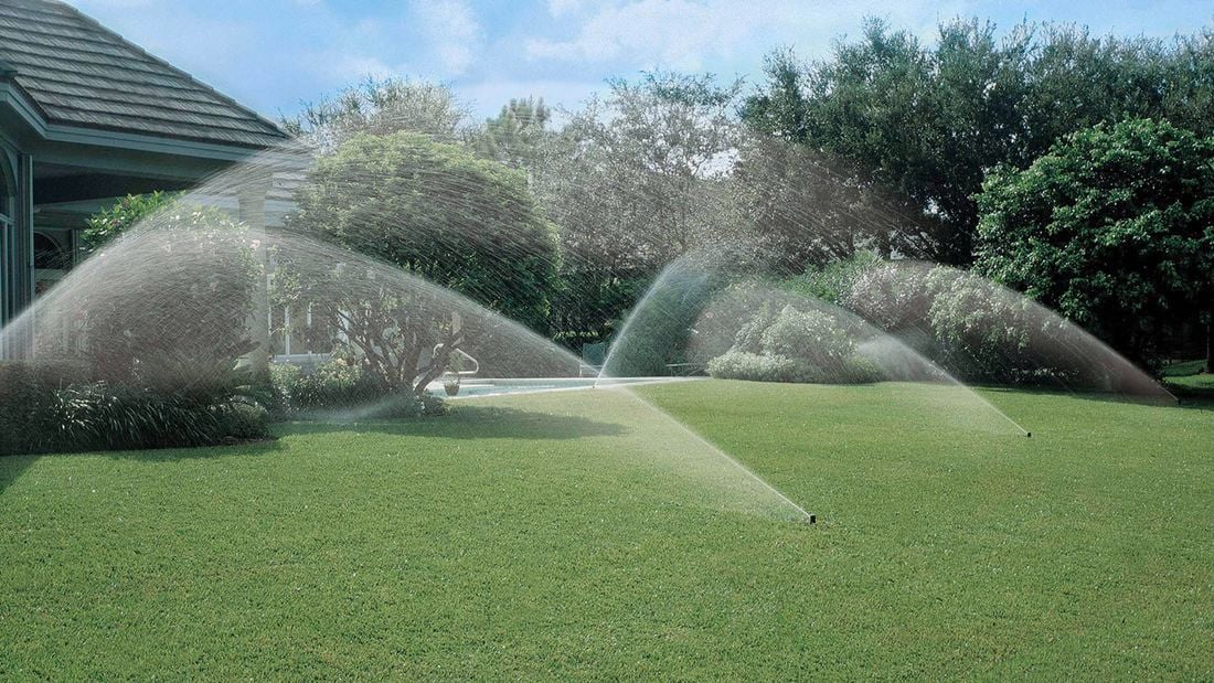 The backyard of a home showing lush green grass with five working irrigation sprinklers