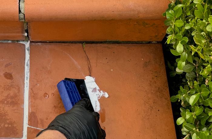 A hand wearing a glove is using squeegee to fill a crack in a tile with exterior grout