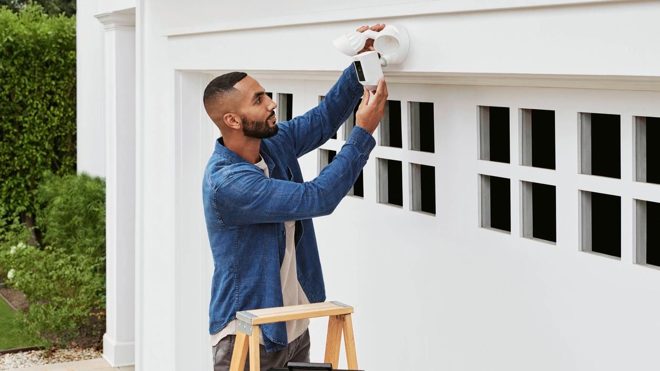 A man on a ladder installing a security camera outside a white-painted garage.