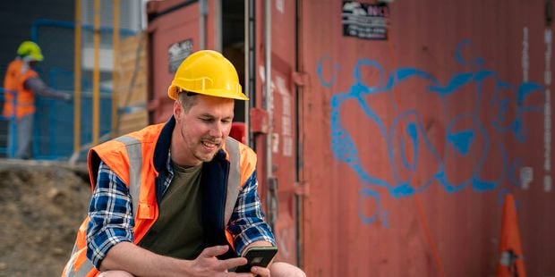 Construction worker in high-visibility vest and yellow hard hat sitting beside a shipping container, using a smartphone, with another worker and scaffolding in the background.