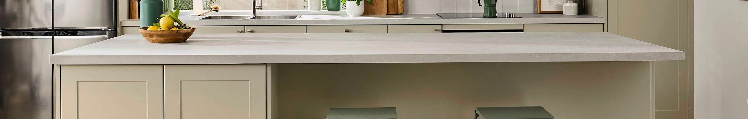 Beige kitchen with a white marble benchtop, two green stools at the bench, with multiple vases with flowers in them on the benchtop.
