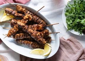  A white platter piled with chipotle chicken skewers with a bowl of rocket leaves on the side