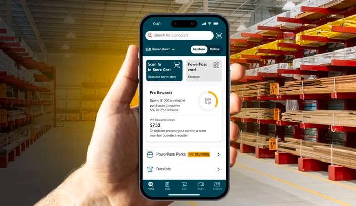 Promotional image showing a hand holding a smartphone displaying a retail hardware app. The phone is centered in front of a large warehouse aisle filled with timber and building materials on industrial shelving, highlighting app features such as rewards, receipts, and in-store shopping tools.