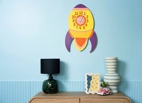 A colourful retro rocket wall clock displayed on a blue wall above a wooden console table with modern decorative pieces.