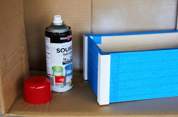  A wooden crate with painters tape attached, inside a sideways cardboard box. An uncapped can of spray paint is next to it.
