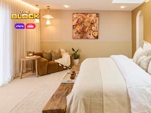 A stylish bedroom featuring a large bed with multiple pillows and a beige color palette. A wooden bench sits at the foot of the bed. To the left, there’s a cozy seating area with a brown sofa, cushions, and two small tables. Large windows with sheer white curtains let in natural light. The walls are decorated with artwork and lighting fixtures. 