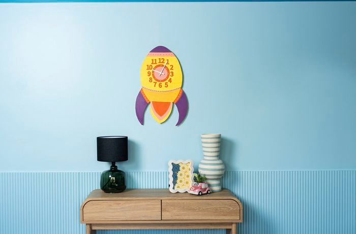 colourful DIY retro rocket wall clock mounted on a blue wall above a wooden console table.