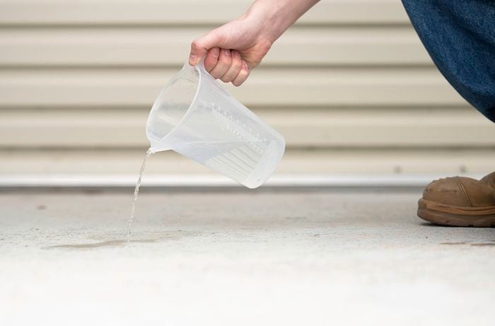 A person is pouring water from a measuring cup onto a concrete surface to check whether the concrete has been previously sealed or coated.