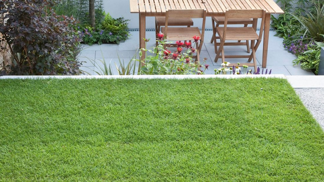 A patch of neat and lush green grass in the foreground. Behind the grass is a sunken grey stone patio with a wooden table and chairs and colourful flowers edging the patio borders