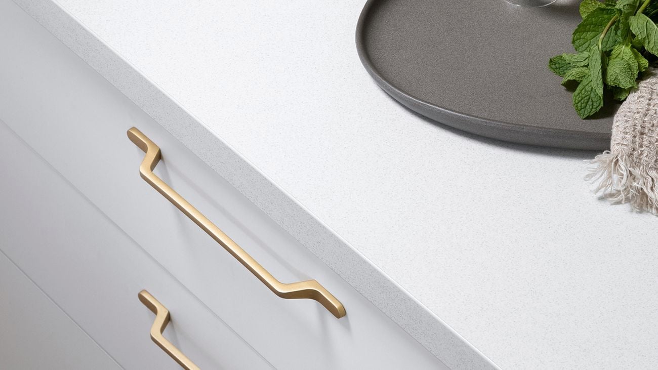 White kitchen countertop with gold drawer handles. On top of the table sits a white stand of fresh lemons, a grey platter and a clear drink with mint.
