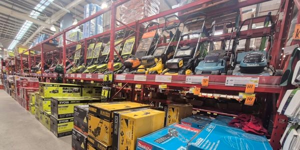Garden equipment aisle at Bunnings Warehouse displaying a range of lawn mowers on elevated shelving and boxed outdoor power equipment from Ryobi, DeWalt, AEG and Makita. Products are arranged beneath warehouse racking with price labels visible along the shelves.