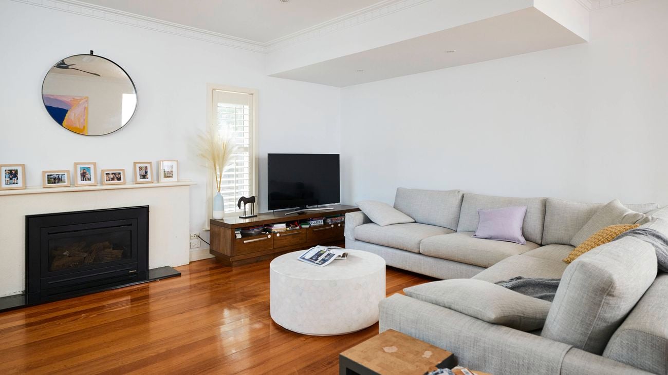Spacious living room with polished wooden floors, a light grey sectional sofa around a round white coffee table, a TV on a low timber unit, and a white fireplace with framed photos and a round mirror, complemented by indoor plants and natural light.