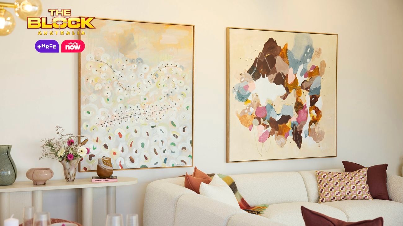 A modern living room featuring a white sectional sofa, a round coffee table, and a built-in fireplace. Two large abstract paintings hang above a console table with decorative items. The room has light-colored walls and wooden flooring. 