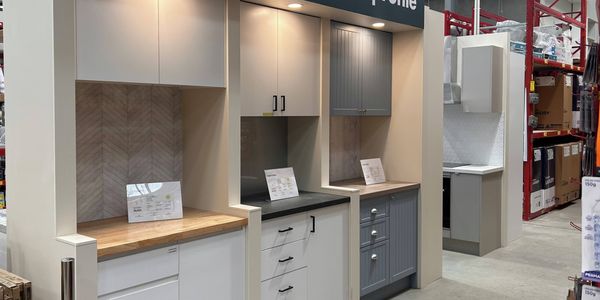 Kitchen cabinetry display area inside a hardware warehouse, showcasing multiple cabinet door styles, benchtops and storage solutions beneath a large sign reading “Choose your perfect profile”. Display units feature a range of colours, finishes and design options.