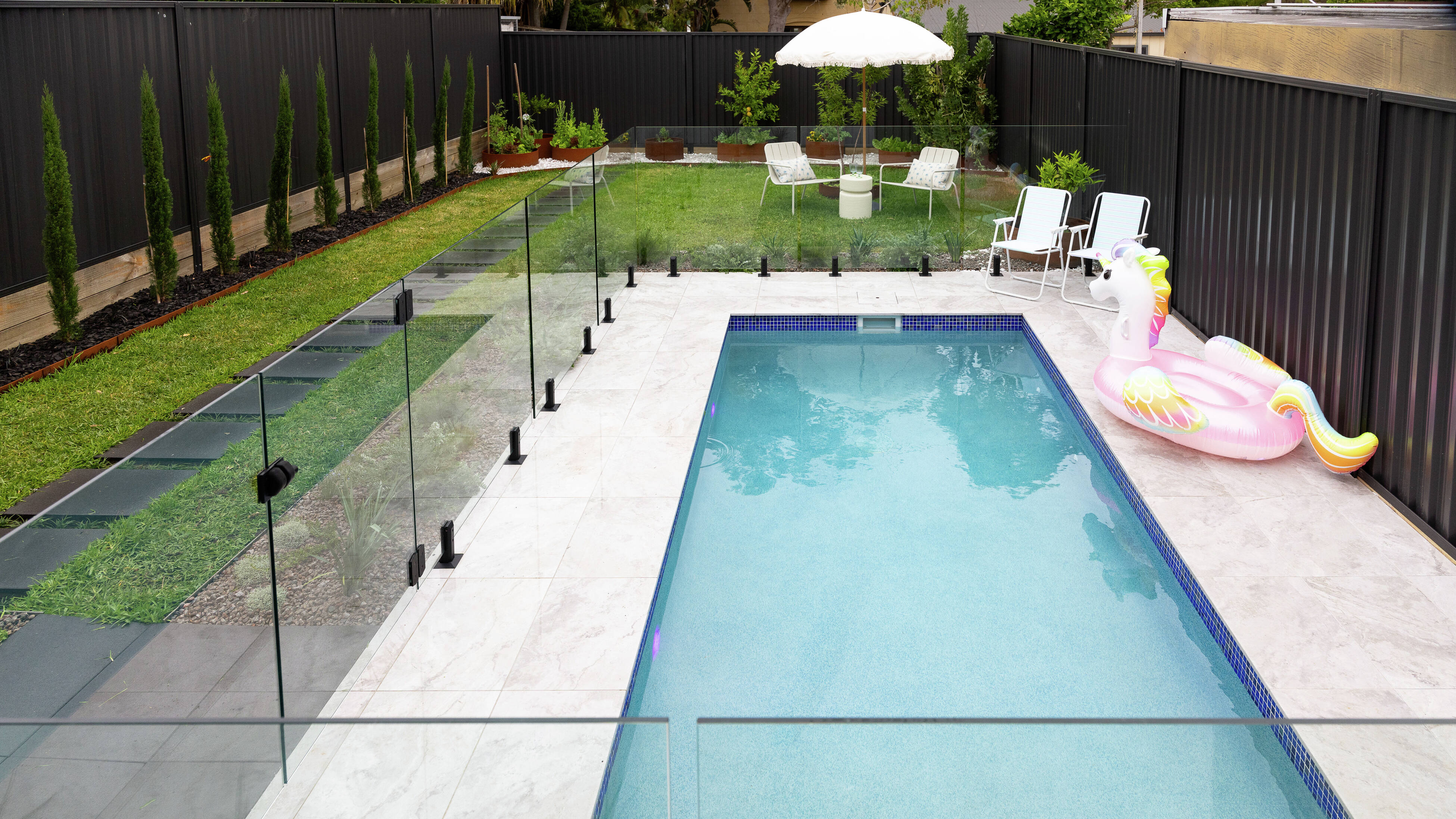 Backyard with a glass-fenced swimming pool, chairs, and umbrella.