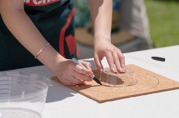 Person using round plastic container to draw circle on cork material.