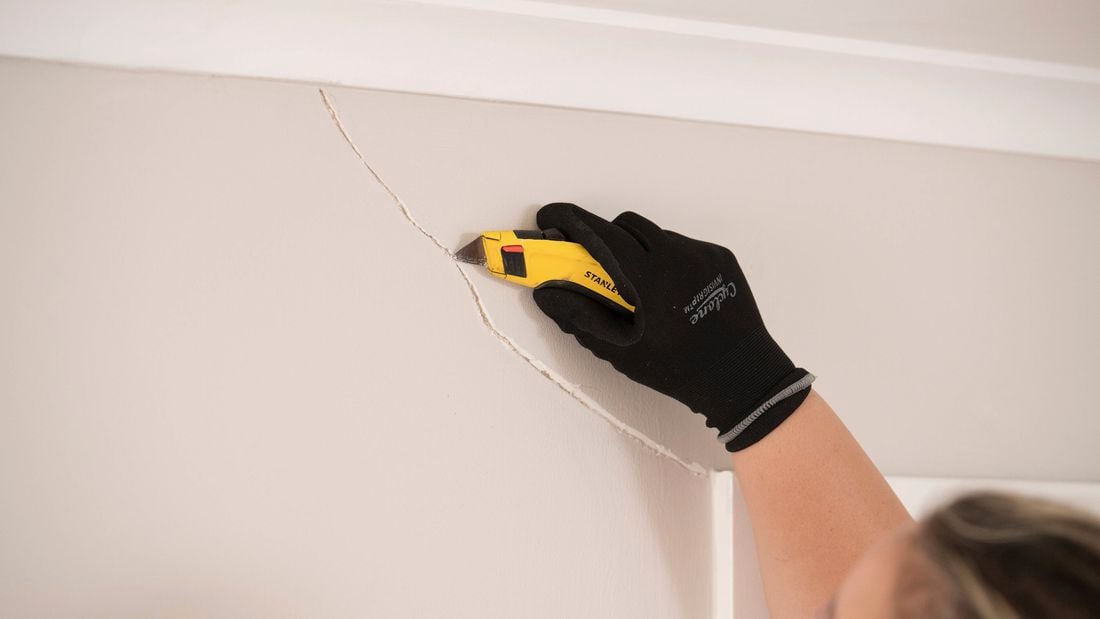 A hand wearing a black safety glove uses a utility knife to repair a cracked wall surface.