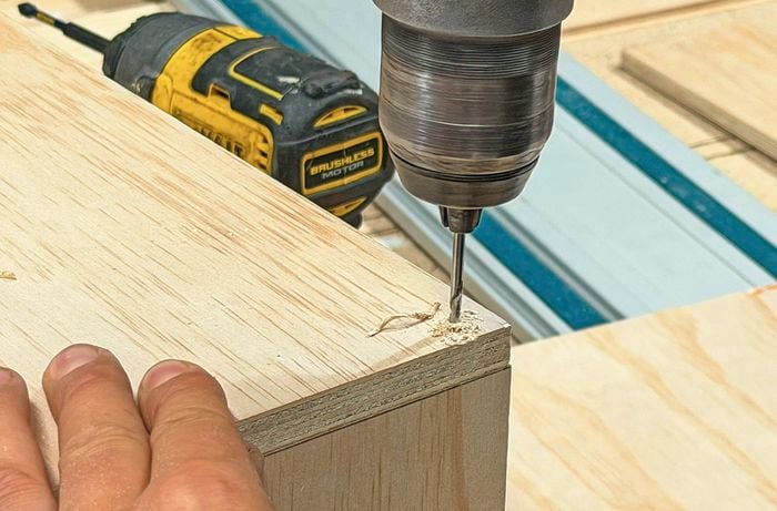 Hands holding a piece of wood and a drill creating a hole in the corner.