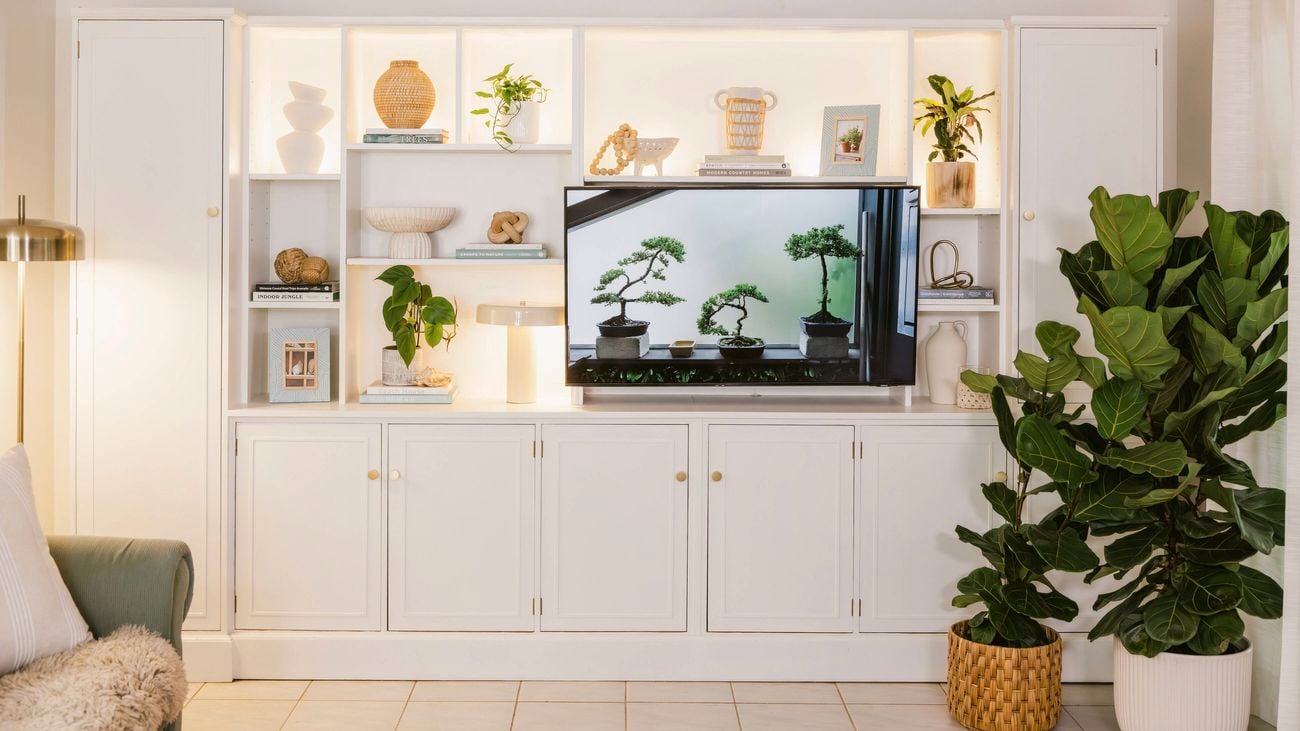 White built‑in living room cabinetry with open shelving, indoor plants, and decorative objects framing a wall‑mounted television