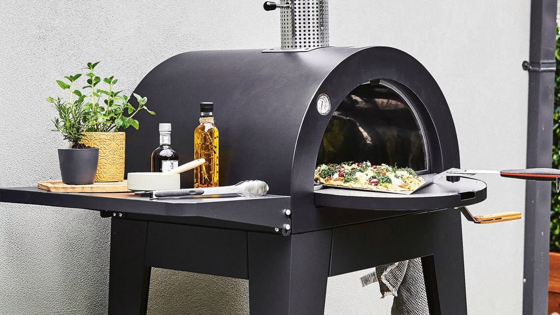 A Bunnings pizza oven with the door opened showing a cooked pizza on a pizza peel being removed.