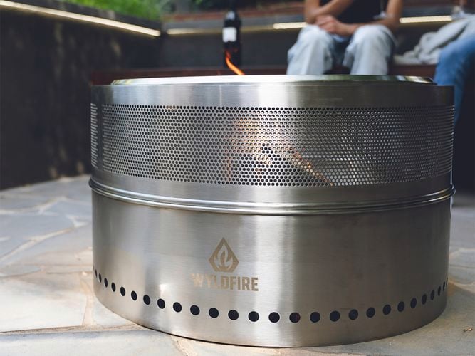 Round stainless steel Wildfire fire pit with visible flame, positioned on an outdoor patio while two people sit behind holding wine glasses.
