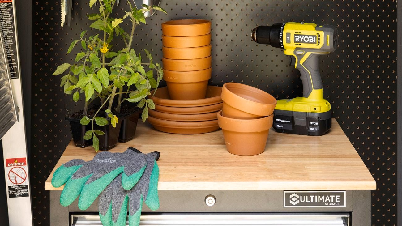 Close up of the interior of a potting shed with a pegboard background, and a drawer unit with a drill, gloves, potted plants and terracotta pots on top.