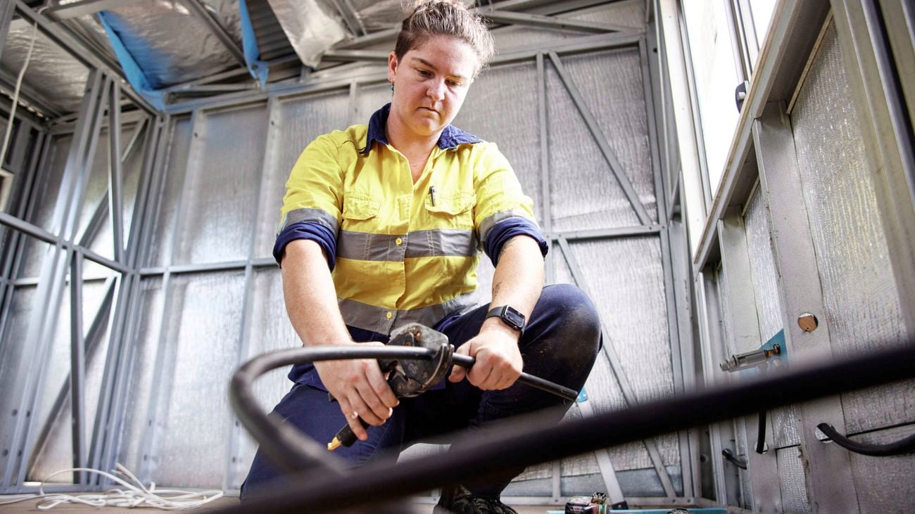 A tradesperson in a hi-vis yellow and navy shirt kneels inside a metal‑framed room, using a tool on an electrical cable