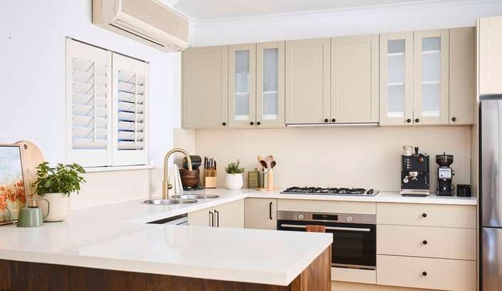 Bright modern kitchen with cream cabinetry, white benchtops, stainless steel appliances, timber island bench and a wall-mounted split-system air conditioner.