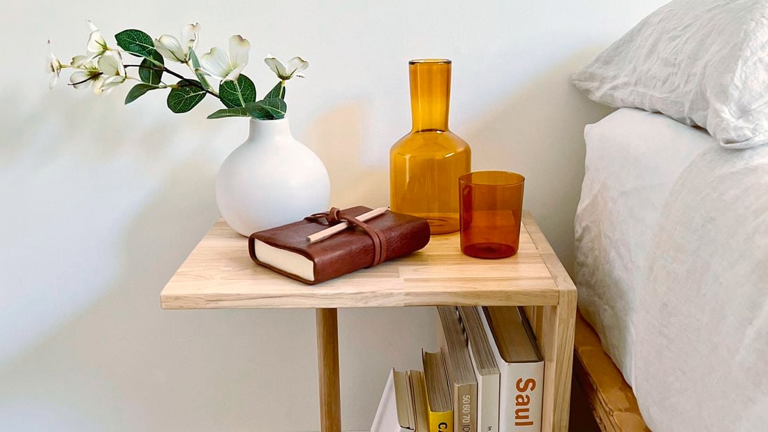 A side table next to a bed with a vase, book, carafe and glass on it