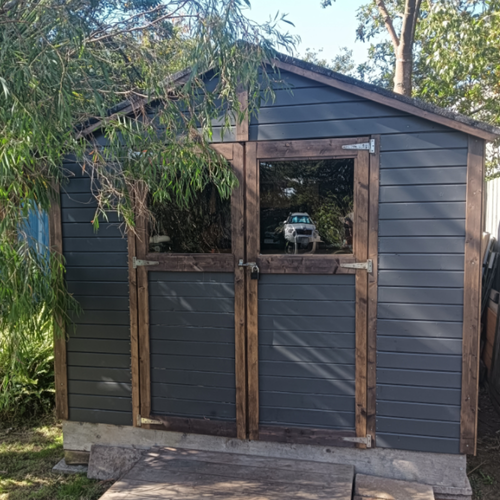 Dark charcoal garden shed with timber trim, pitched roof, and double doors with large windows, set among mature trees and garden planting.