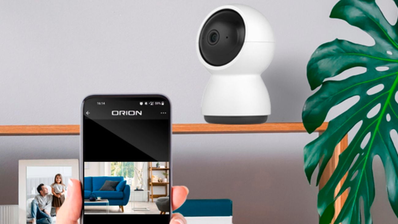 Living room with wooden shelf display, holding picture frames and a home security camera. A hand holding a smartphone with a picture of a blue sofa on the screen.