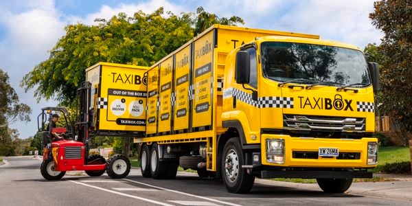 Yellow TAXIBOX truck parked on a suburban street with multiple storage boxes loaded on the back, while a small red forklift lifts another yellow TAXIBOX unit beside the truck. Trees and houses are visible in the background under a partly cloudy sky.