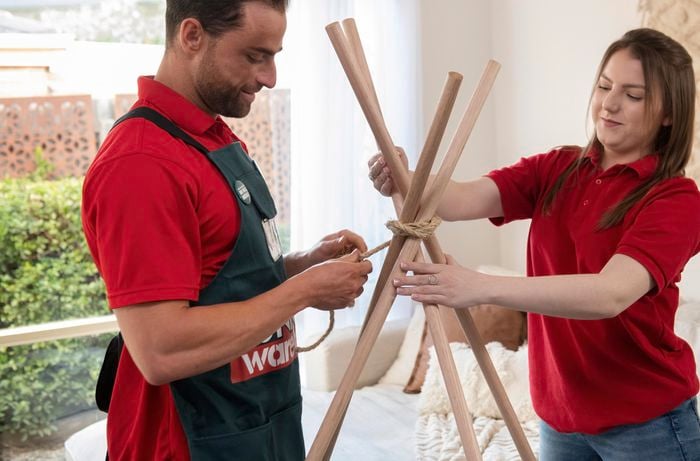 Person holding three bits of wood and another person tying rope around it.