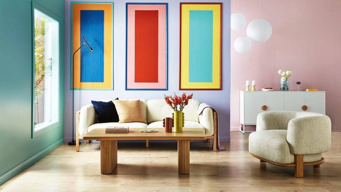 Living room with a cream colour sofa, three colourful rectangular painted wall panels on a purple wall, a tan coffee table and a cream colour bean shaped sofa chair