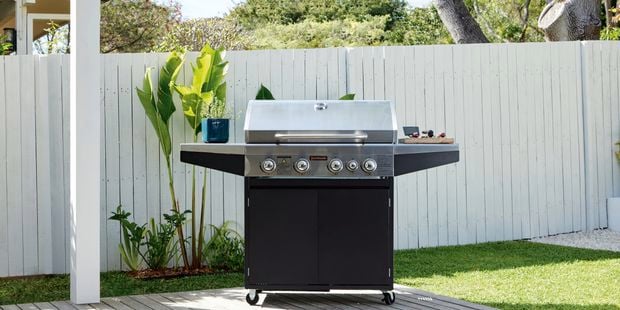 Barbecue sitting on deck with lid closed, white fence and plants are seen in the background.