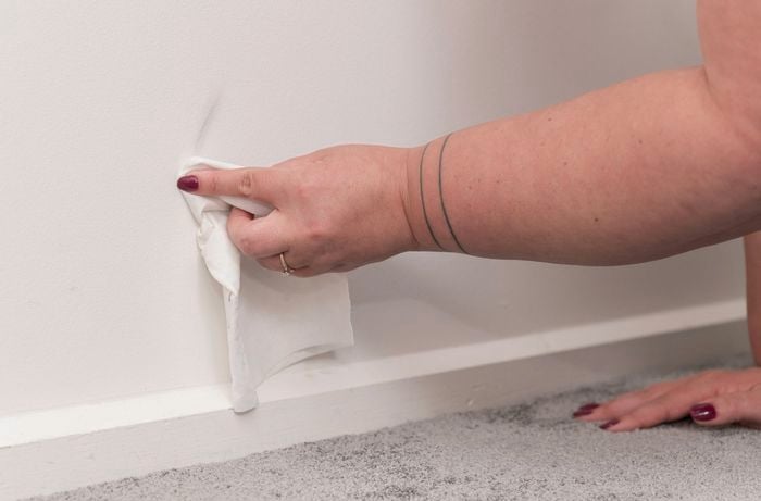 A person is cleaning a stain off a white wall with a sugar soap wipe while their other hand rests on a carpeted floor. 
