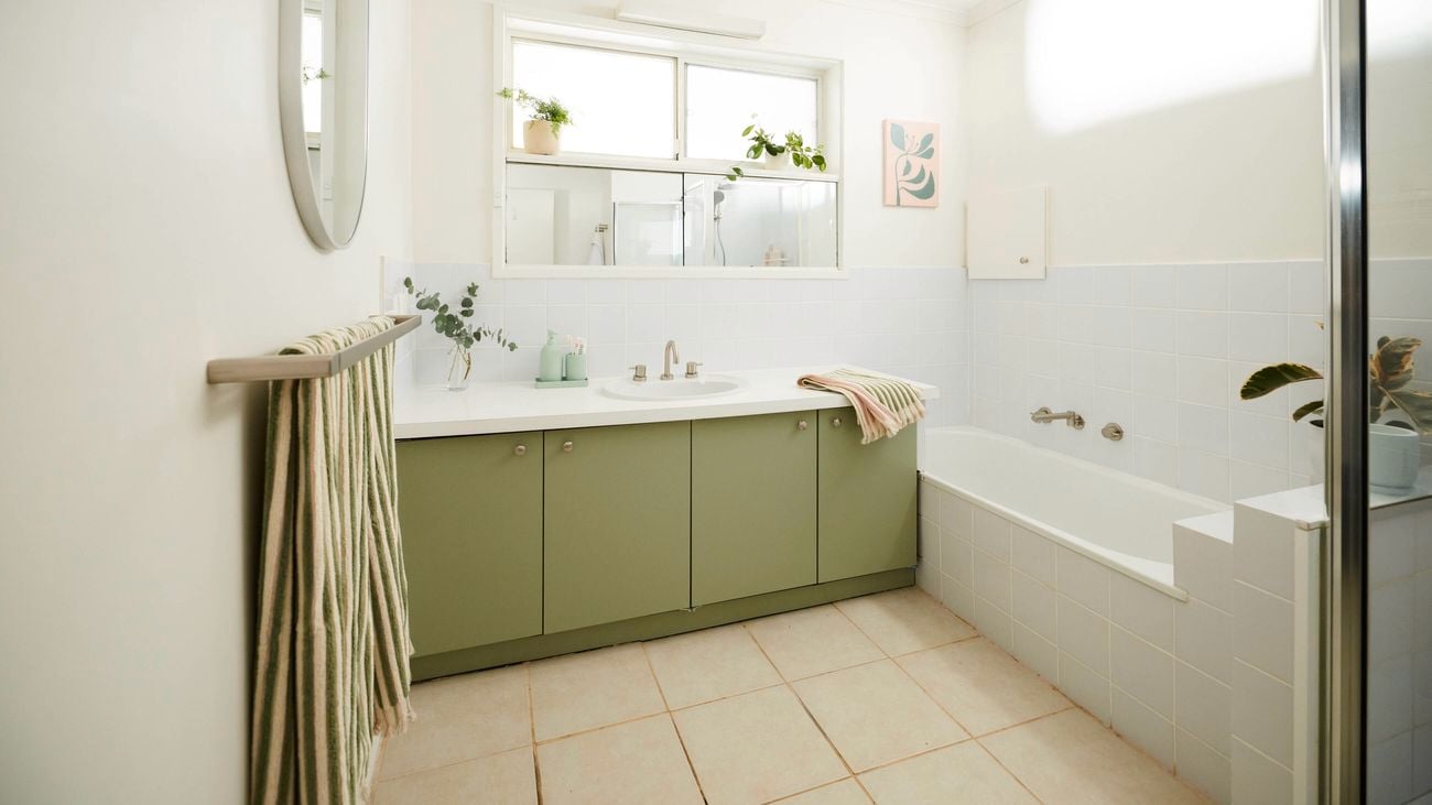 Bright bathroom with a green vanity cabinet and white benchtop, featuring a central sink beneath a wide mirror, a bathtub to the side, white tiled walls, striped towel on a rack, and potted plants along the windowsill for natural light and greenery.