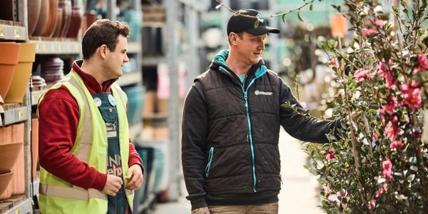 A Bunnings worker in a high-visibility vest talks with a customer while looking at flowering plants in a garden center aisle lined with pots.