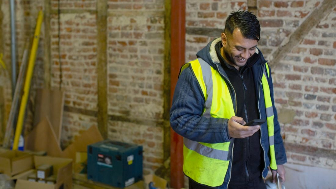 A tradesperson in a high-vis vest looking at his phone on a construction site.